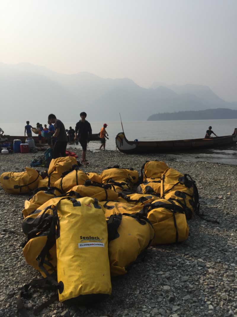 Welded waterproof camping bags in use at a campsite near water — roll-top backpack, duffel, and dry sack