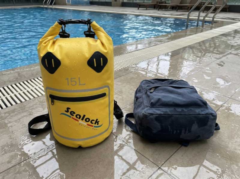 PVC-based wet dry bag on a wet pool deck beside a fabric bag with a soaked base